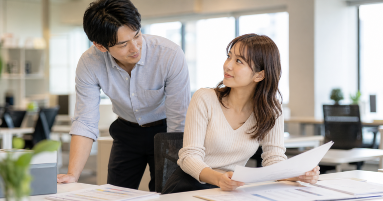 職場でデスクの後ろに少し離れて立ち、資料をのぞきこむ男性と、気づいた様子で振り返る女性の写真