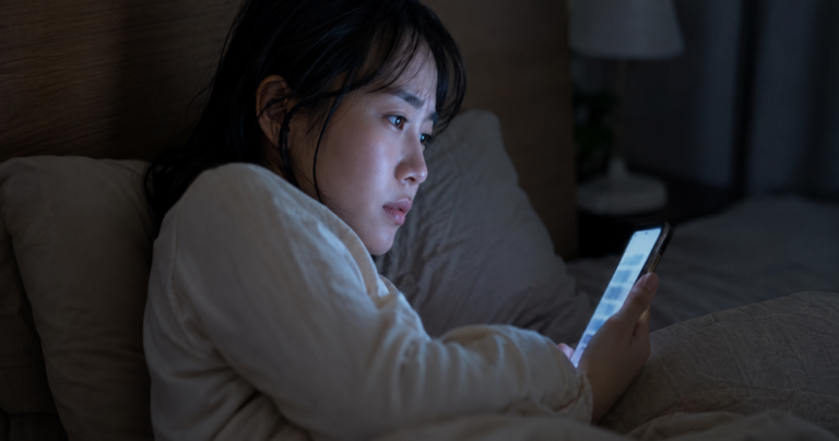 夜、ベッドの上でスマートフォンの画面を見つめながら不安そうな表情を浮かべる女性の写真
