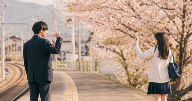 桜の季節に、進学で離れ離れになる高校生カップルが最後に手を振り合っている後ろ姿の写真