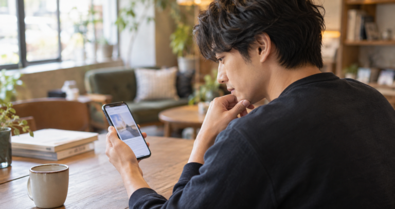 スマートフォンの画面を見ながら少し考え込むような表情を浮かべる、落ち着いた雰囲気の男性の写真