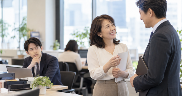 オフィスで年上の女性社員が甲高い声で愛嬌を振る舞い、周囲の同僚が戸惑っている様子のイラスト