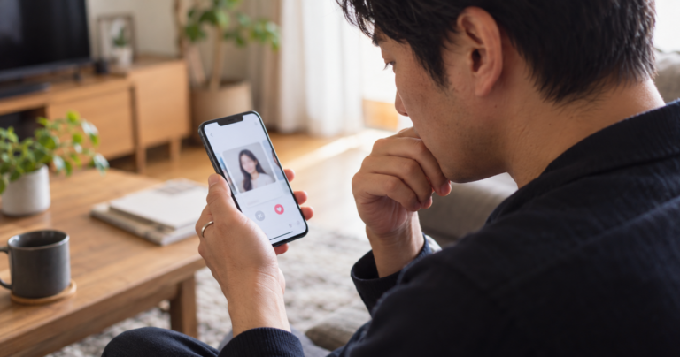 スマートフォンでマッチングアプリの画面を見つめながら、結婚指輪をした手で考え込む男性の写真
